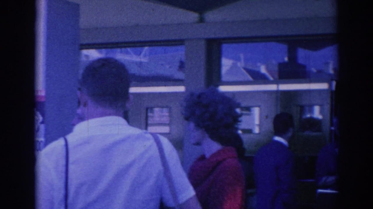 1967 Los Angeles street frame with adults waiting near a building, a woman in red, a man in a white shirt, dark suits, and visible wall notices.