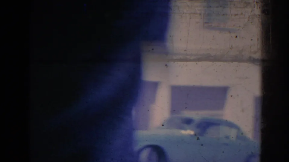 1967 Los Angeles home-movie frame with a blurred car moving past a building, heavy gate shadow, blue cast, and visible film wear.