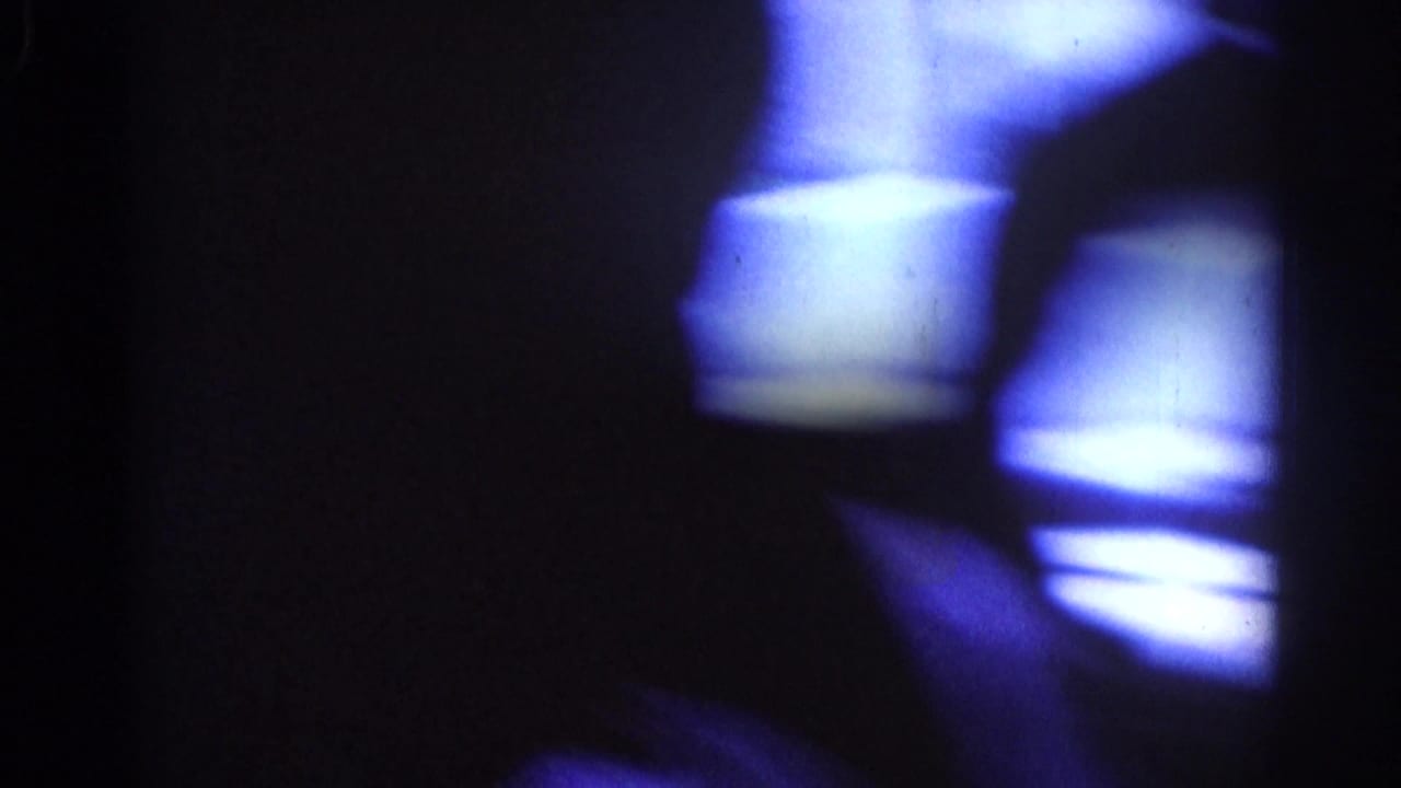 Dark purple-tinted blurred frame with partial light shapes and film marks.