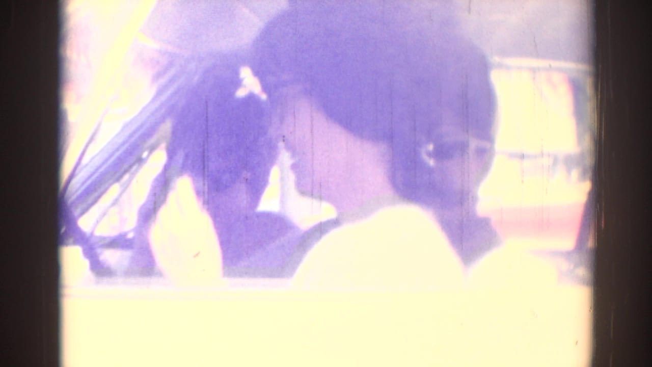 Two passengers inside a car in the 1967 Los Angeles home movie, seen through bright window glare and a purple color cast.