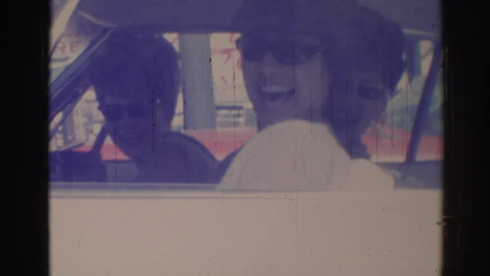 Two people seated in a car in 1967 Los Angeles, one in sunglasses, looking toward the home-movie camera through soft focus.