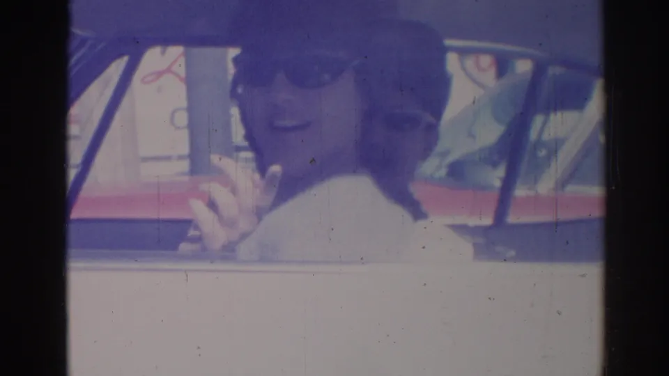 A sunglasses-wearing passenger smiles and gestures toward the camera beside another passenger in the 1967 Los Angeles car scene.