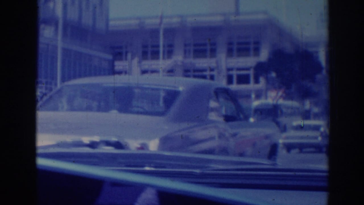 Street view through a car windshield with parked mid-century cars and a building beyond.