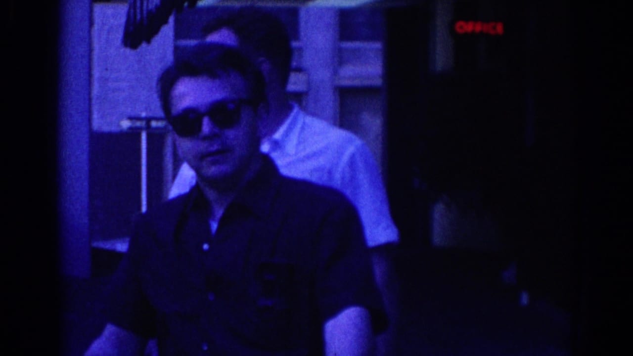 Adult man in sunglasses and dark shirt near a building entrance in 1967 Los Angeles, with a second man in a white shirt partly visible.