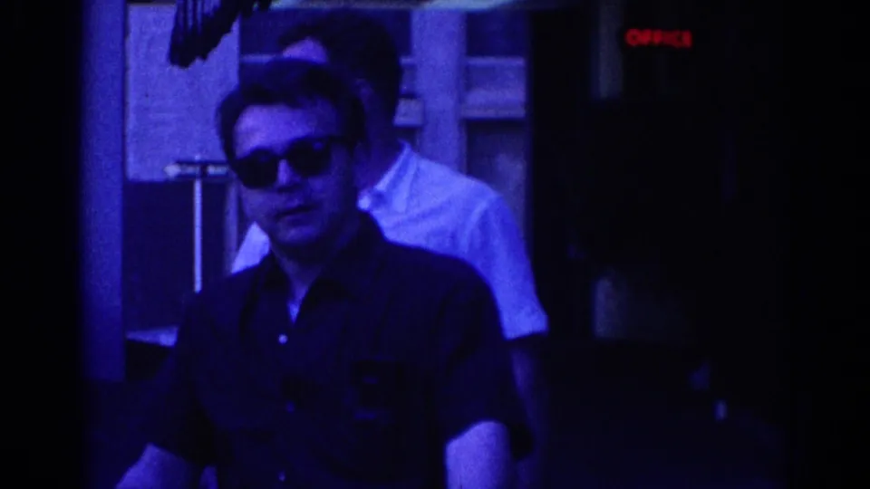 Adult man in sunglasses and dark shirt near a building entrance in 1967 Los Angeles, with a second man in a white shirt partly visible.