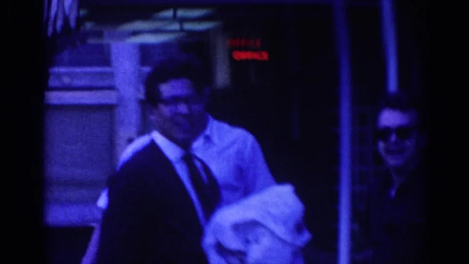 Adult man in white shirt and tie holding a light garment outside an office area, with another sunglassed man partly visible beside him.