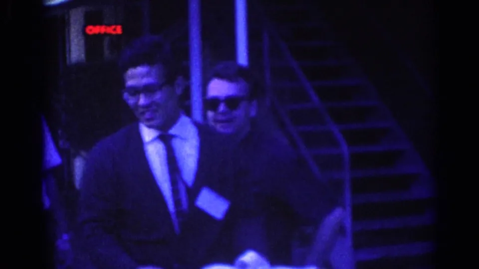 Two adult men in suits near an exterior stairway in Los Angeles, with sunglasses, ties, papers or a small object, and a red OFFICE sign visible.