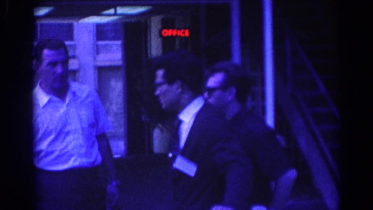 Three men stand outside a building near a glowing red OFFICE sign.