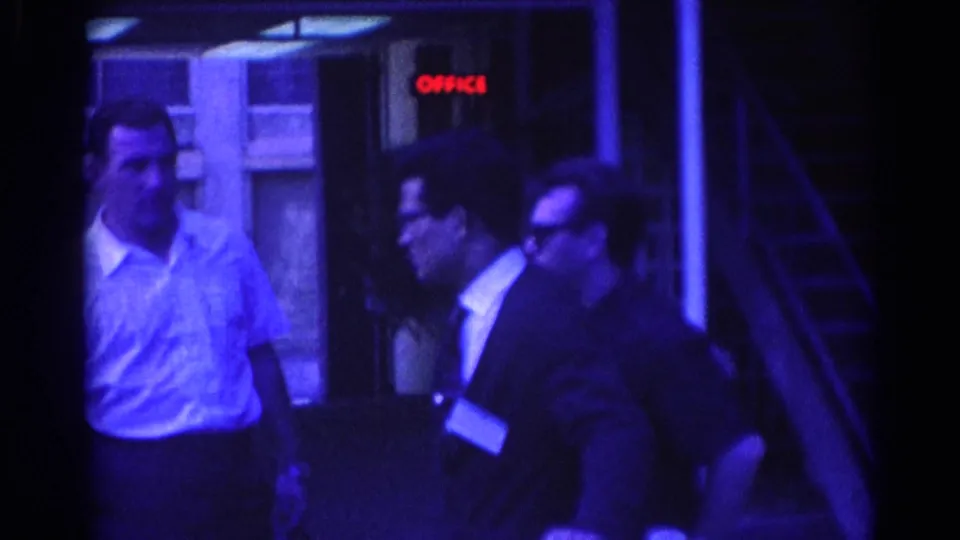 1967 Los Angeles home-movie frame of well-dressed men near windows and a red OFFICE sign, with blue-purple film tint.