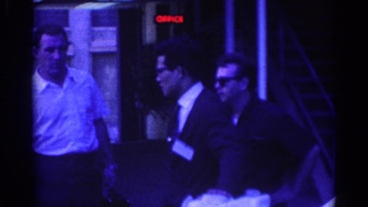 Well-dressed men are filmed outside a Los Angeles building in 1967, with one man turned toward another near a red OFFICE sign.