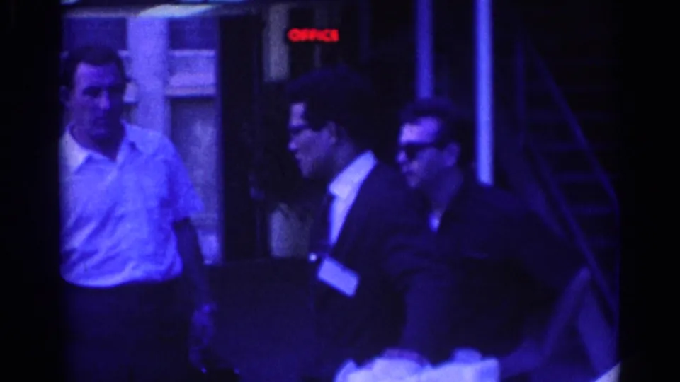 Well-dressed men are filmed outside a Los Angeles building in 1967, with one man turned toward another near a red OFFICE sign.