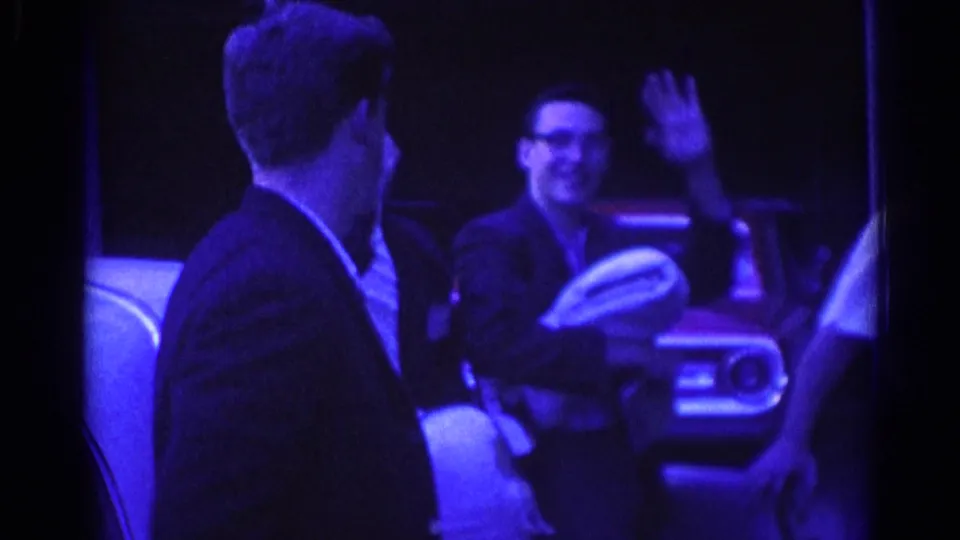 A man in glasses appears to wave while holding a light cloth or hat near other men and a parked car in 1967 Los Angeles footage.