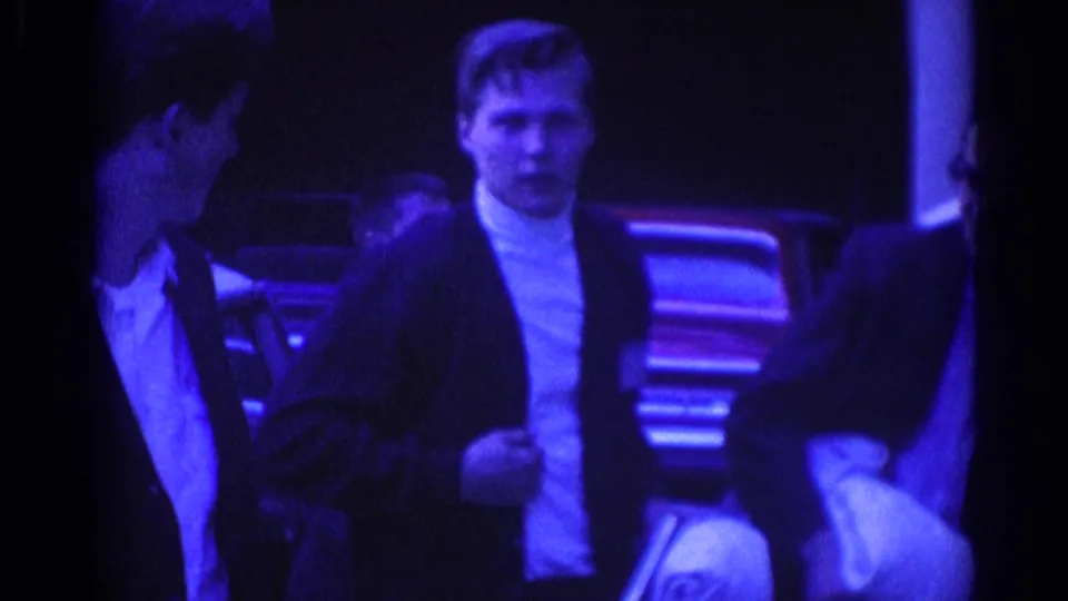 A well-dressed man in a dark jacket and white turtleneck stands with other men beside a parked car in blue-purple 8mm footage.