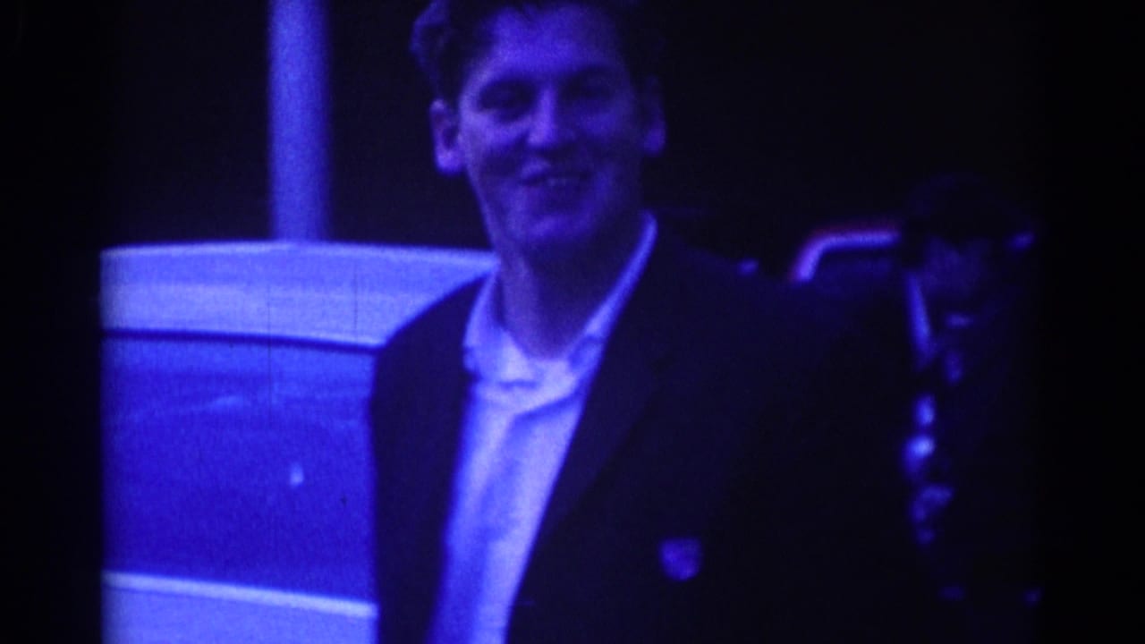 Smiling man in a dark suit near a parked car at night.