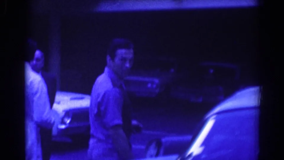 A man in a short-sleeve shirt looks toward the camera near parked cars in the 1967 Los Angeles night footage.