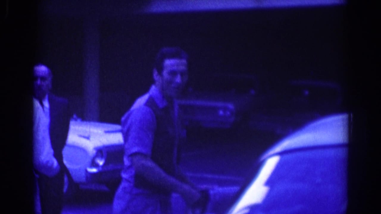 Man in a dark vest walks near a parked car at night while another man stands behind him.