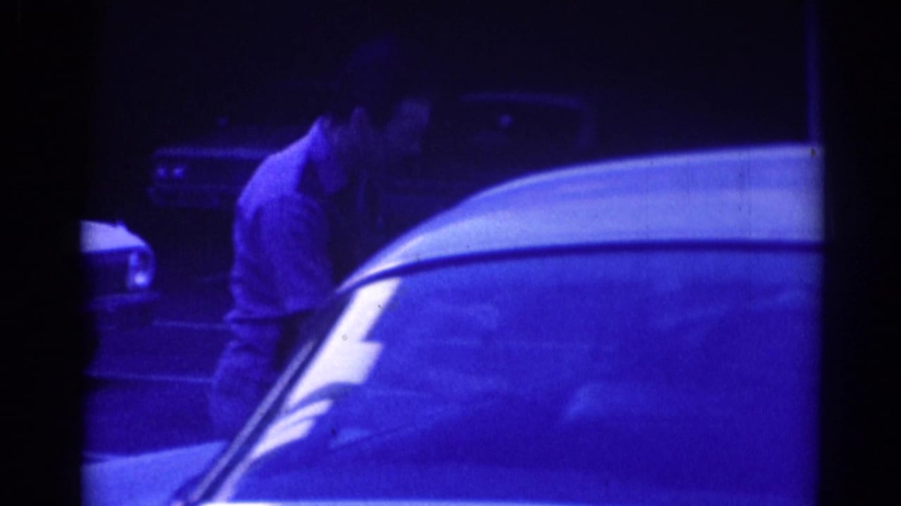 Blue-tinted 1967 Los Angeles home-movie frame of a man reaching or leaning into a parked car window at night.