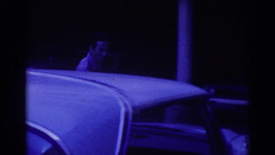 Low-light 8mm frame from 1967 Los Angeles with a parked car roof in front and a partially seen man behind it.