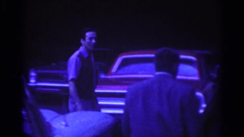 1967 Los Angeles night frame showing a man beside parked classic cars, with another figure seen from behind in the foreground.