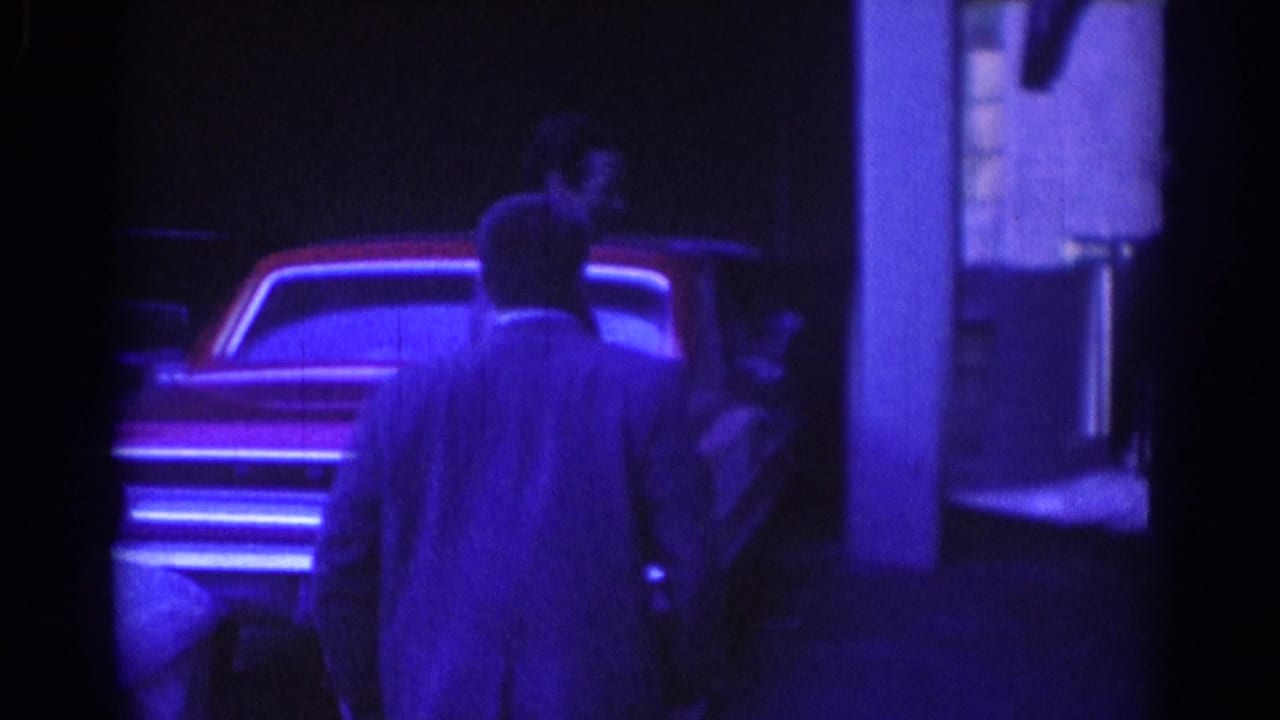 Man seen from behind near a parked car and building entrance in blue-tinted low light.
