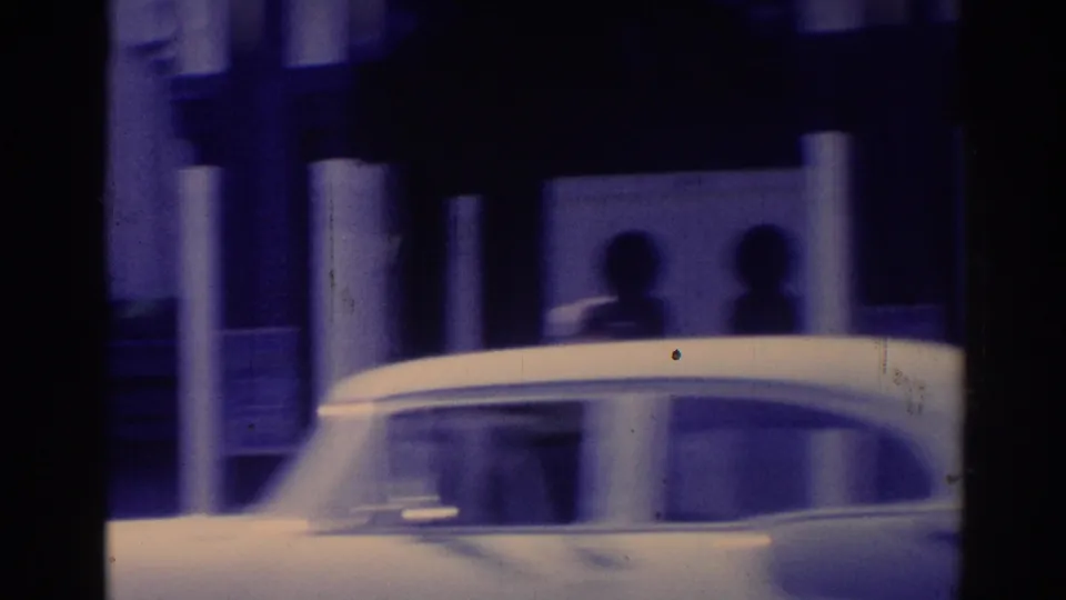 A light-colored car passes close to the camera, with building columns and shadowed people behind it.