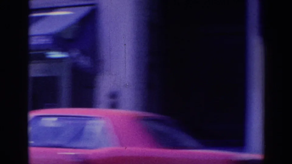 A red car crosses the foreground against shaded columns, with the handheld 8mm frame partly obscured.