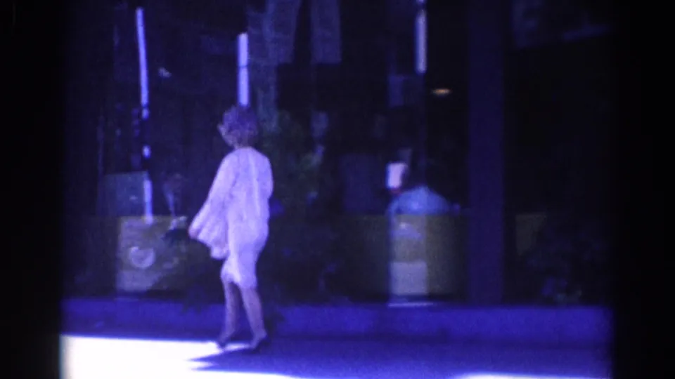 The woman continues across the frame in a light dress and heels, with sidewalk fixtures and shadowed storefronts behind her.