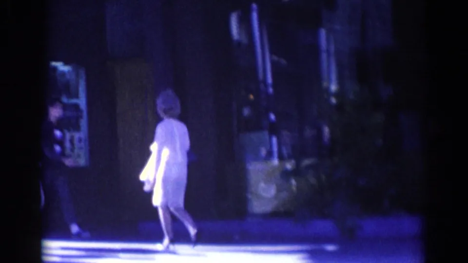 Near the end of the 1967 Los Angeles clip, the woman walks away through bright sidewalk light beside a shadowed building front.