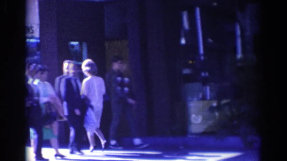 1967 Los Angeles home-movie frame with pedestrians in suits and dresses passing storefronts under deep blue shadow and bright sidewalk light.