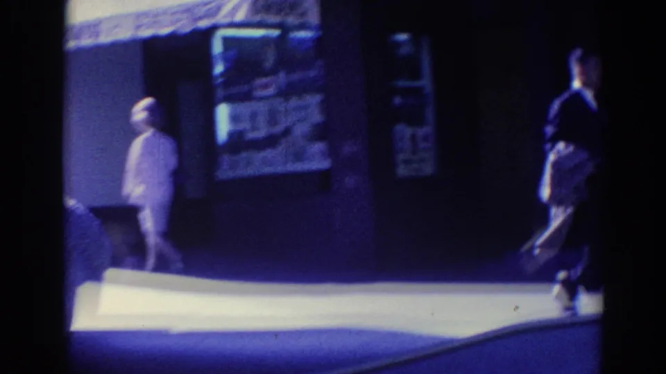 Soft 8mm frame of a Los Angeles sidewalk with a woman in a light outfit near signage and another pedestrian crossing the shaded foreground.