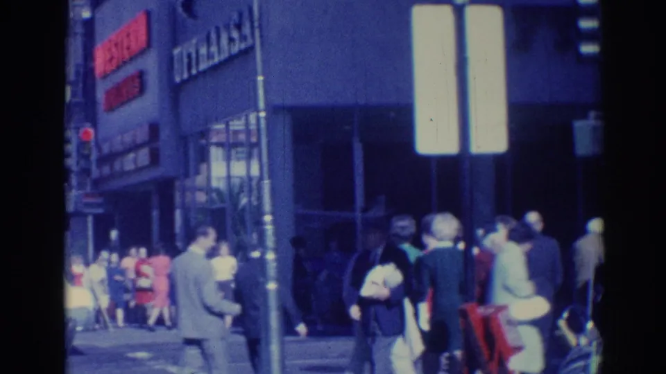 1967 Los Angeles street corner view with pedestrians near a commercial facade, street poles, bright signs, and mid-century clothing.