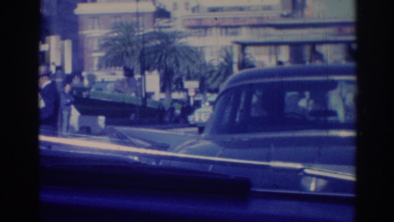 Moving-vehicle view of 1967 Los Angeles traffic with classic cars, pedestrians, palm trees, and large urban buildings.