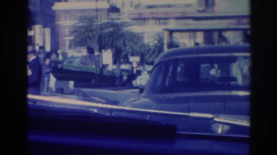 Moving-vehicle view of 1967 Los Angeles traffic with classic cars, pedestrians, palm trees, and large urban buildings.