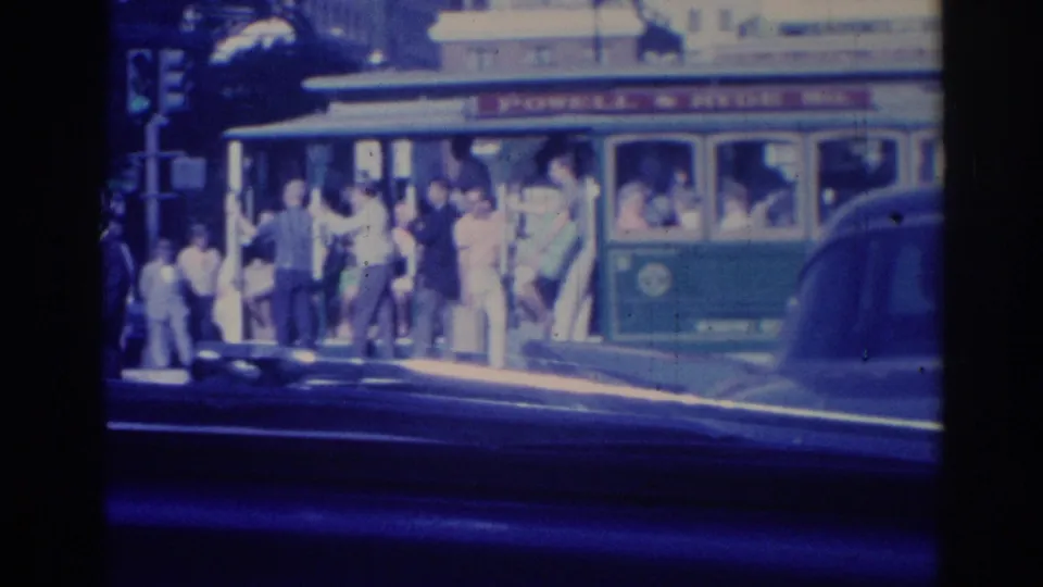 1967 Los Angeles street scene showing a green transit vehicle, sidewalk pedestrians, and classic cars from a moving vantage point.