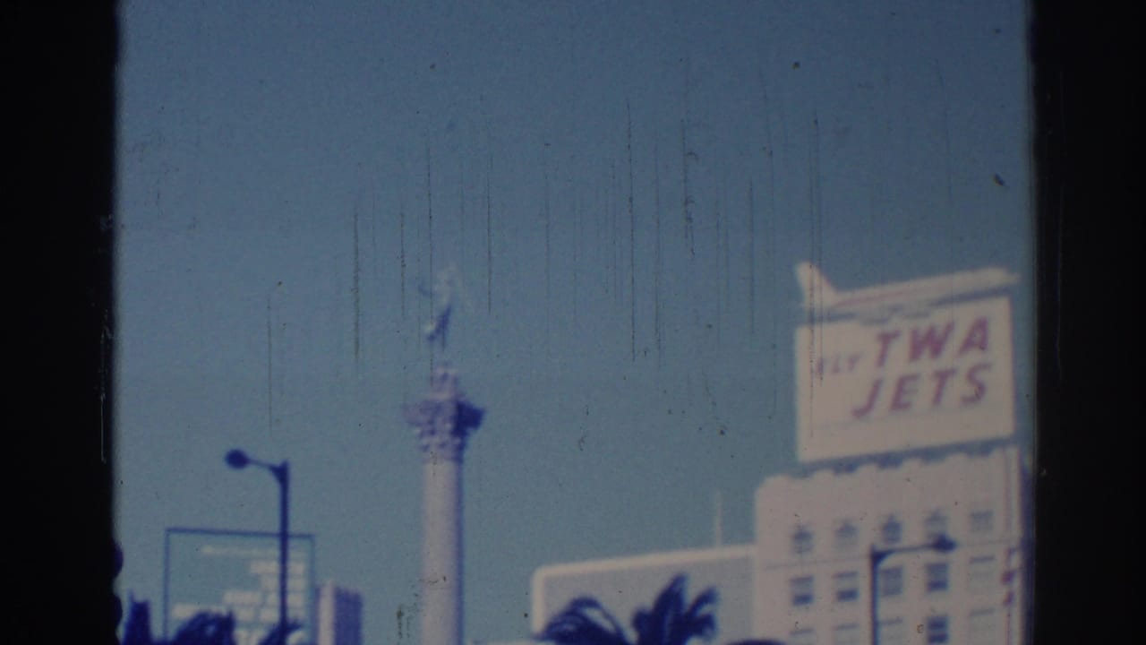 1967 Los Angeles street view with a TWA Jets billboard, a tall tower or monument, lamp posts, and palm tops under clear sky.