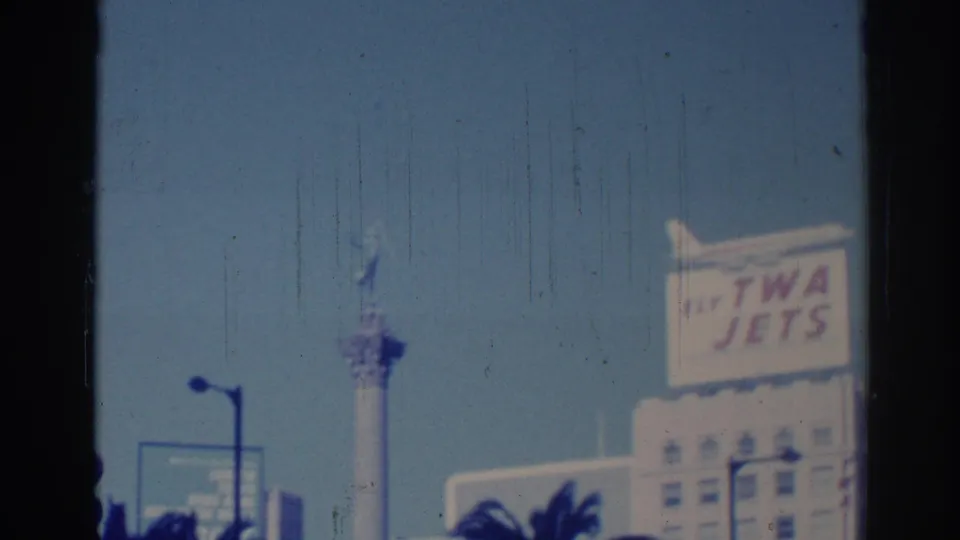 1967 Los Angeles street view with a TWA Jets billboard, a tall tower or monument, lamp posts, and palm tops under clear sky.