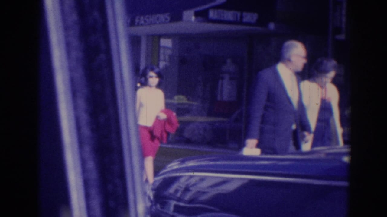 Adults walking near a storefront and dark car on a city sidewalk.