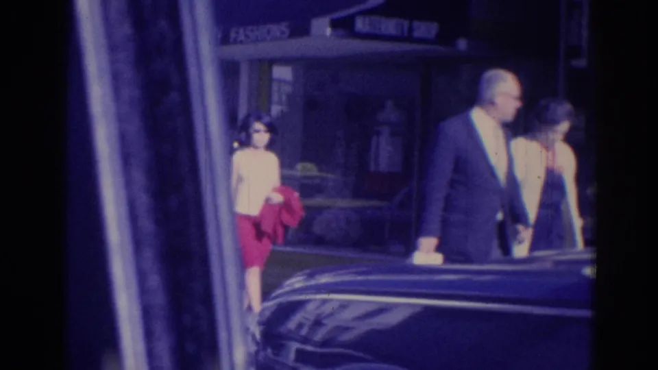 Adults in 1960s attire pass a storefront in Los Angeles, with a dark car in the foreground and shop signs above the entrance.