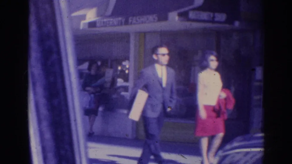 A man in a suit and a woman in a white top and red skirt walk by a Los Angeles storefront marked maternity fashions.