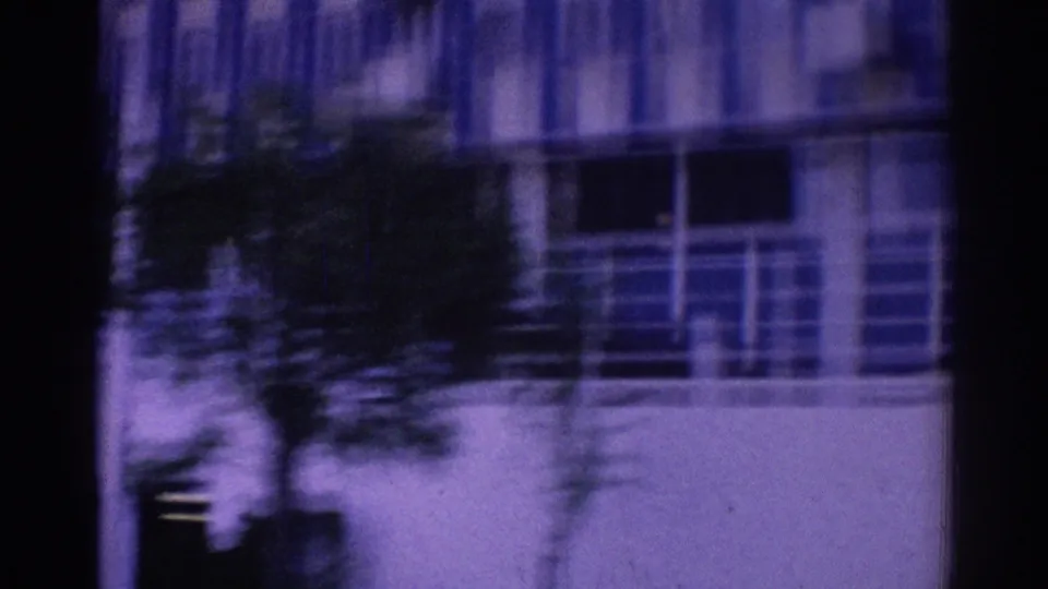 Moving 8mm view of a Los Angeles hotel facade with balcony rails, windows, a tree silhouette, and dark vehicle-window edges.