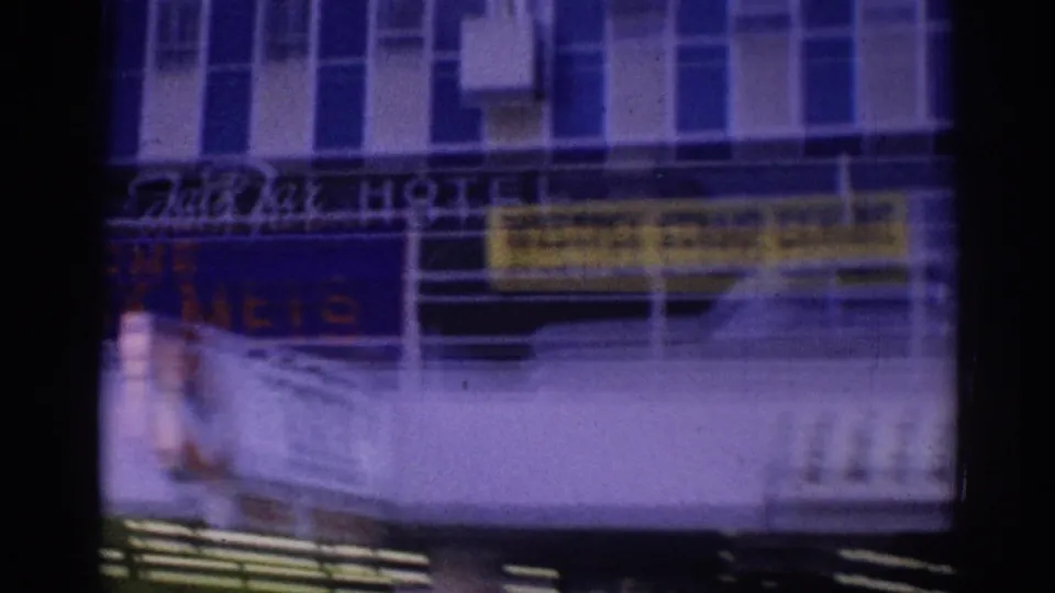 Moving vehicle view of a Los Angeles hotel exterior with visible hotel lettering, yellow banner signage, balconies, and windows.