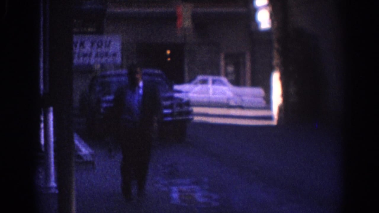 A suited man walks along a dim street near parked cars and signage.