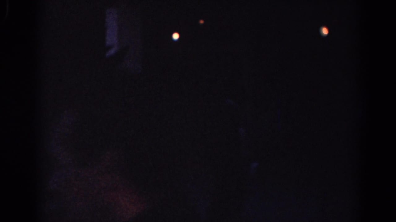 Low-light 1967 Los Angeles home-movie frame with two distant lights and a shadowy figure or shape near a building exterior.