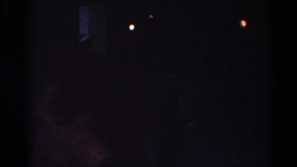 Low-light 1967 Los Angeles home-movie frame with two distant lights and a shadowy figure or shape near a building exterior.