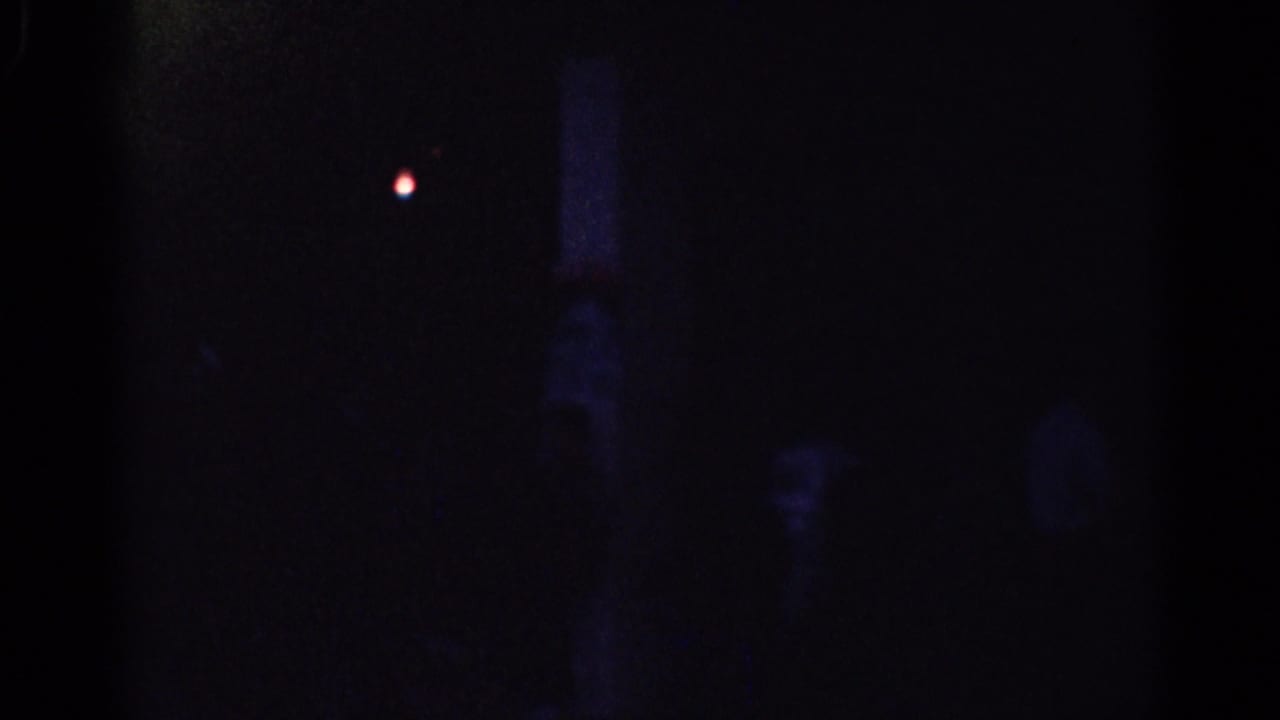 Very dark indoor frame with a small red light and faint human shapes.