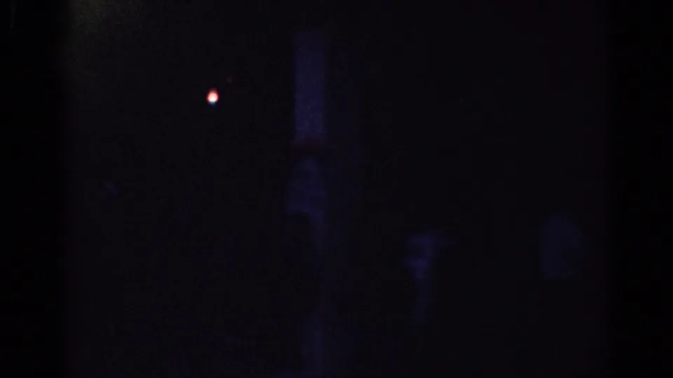 Underexposed 1967 Los Angeles 8mm frame with a small red point, dark glassy surfaces, and barely visible figures in shadow.