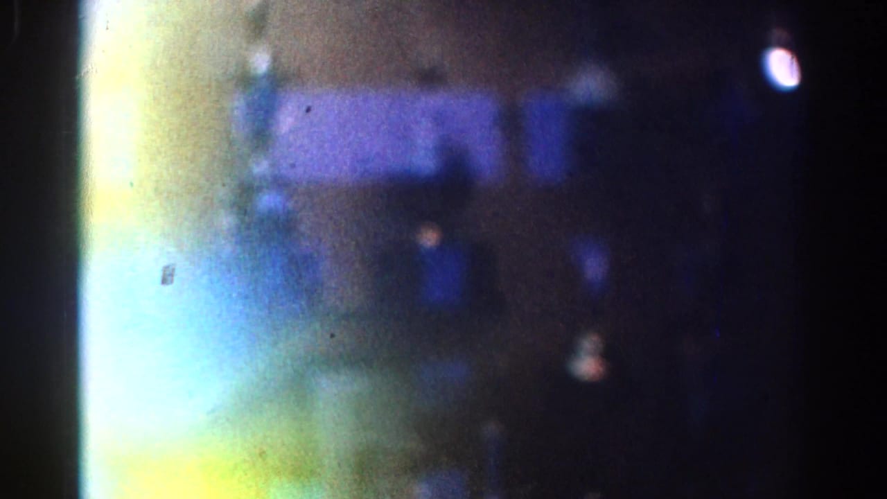 Soft, grainy 1967 Los Angeles frame showing a reflected glass display area with blue bands, yellow-green flare, and a bright white spot.