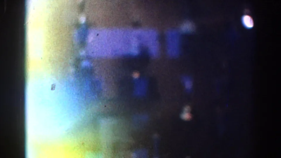 Soft, grainy 1967 Los Angeles frame showing a reflected glass display area with blue bands, yellow-green flare, and a bright white spot.