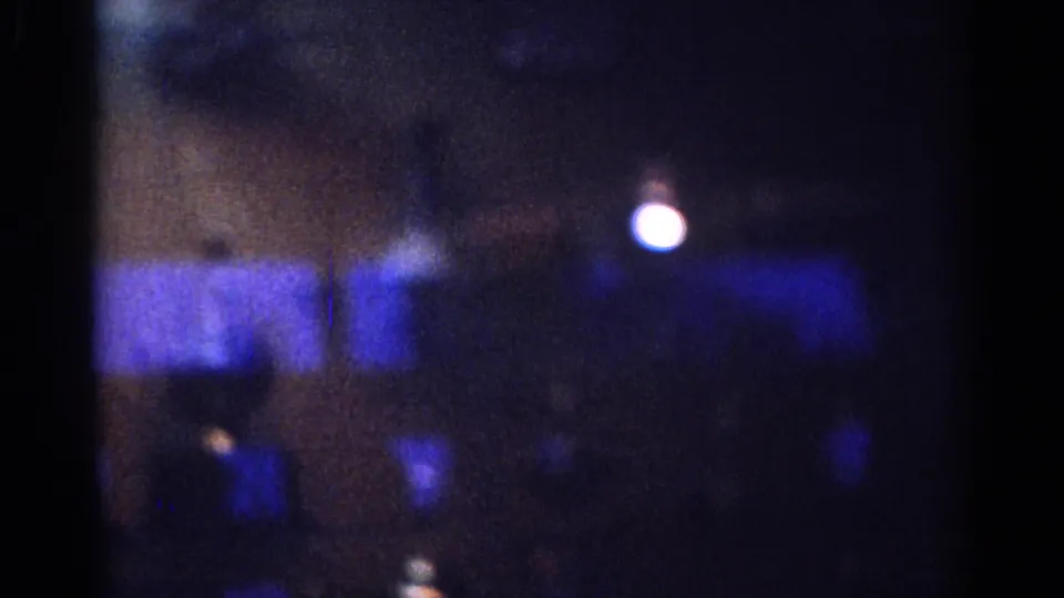 Grainy 1967 Los Angeles home-movie frame with a bright white orb, blue reflected bands, and indistinct display shapes behind glass.
