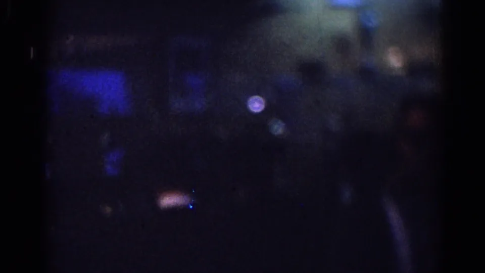 Underexposed 1967 Los Angeles 8mm frame showing blue highlights in a dark glassy scene, a small pale light, and indistinct human forms.